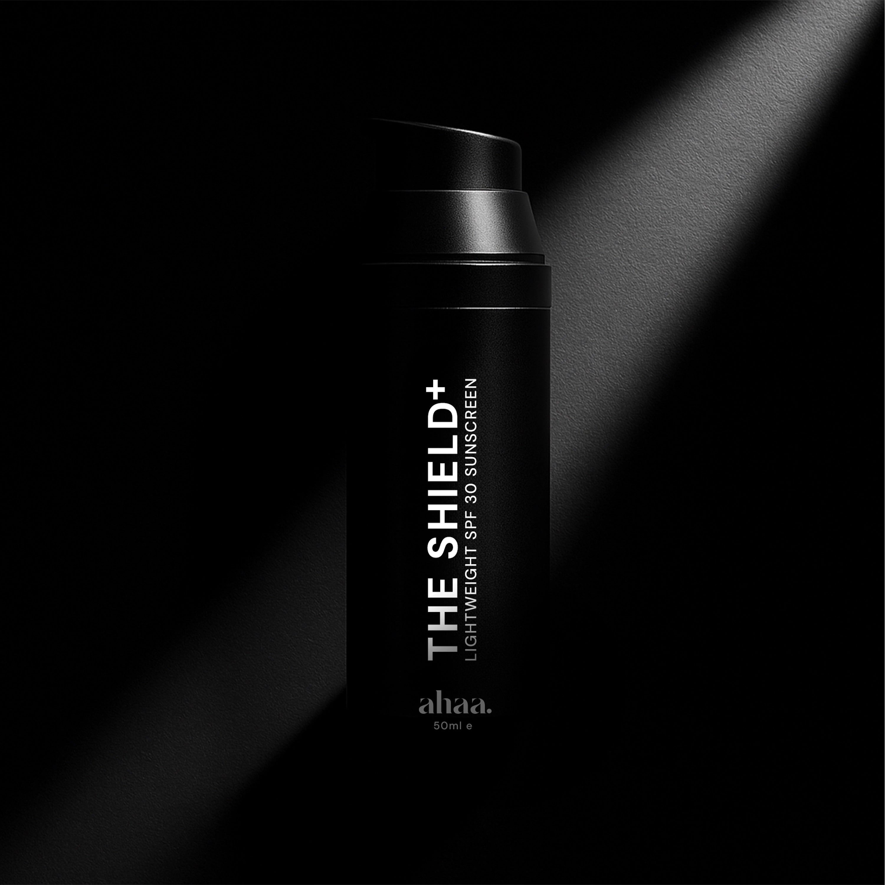 Black sunscreen bottle labeled 'The Shield+' by Ahaa Skincare on a dark background